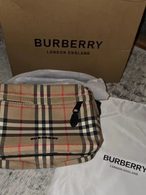 Burberry väska  - Säljer en klassisk rutig axelväska från Burberry i beige, svart, rött och vitt. Väskan har justerbar axelrem, dragkedja och det ikoniska Burberry-mönstret. Perfekt för dig som vill ha en stilren och tidlös accessoar. Den är helt ny, dustbag och box medföljer. Tveka inte för att ställa frågor. 