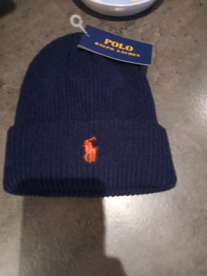 Mössa - Snygg mörkblå ribbstickad mössa med klassisk orange logga broderad framtill. Mössan har uppvikt kant och är tillverkad i mjukt material som håller dig varm och stilren under kalla dagar.