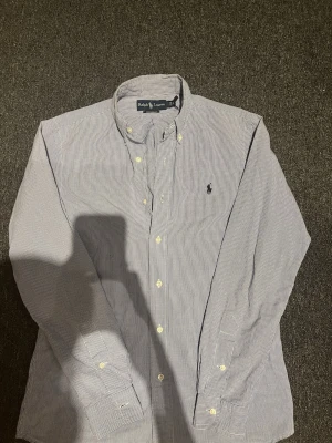 Randig skjorta från Ralph Lauren - Klassisk skjorta från Ralph Lauren med smala blå och vita ränder, button-down krage och broderad logga på bröstet. Skjortan har långa ärmar och vita knappar framtill. Perfekt för en clean och stilren look.