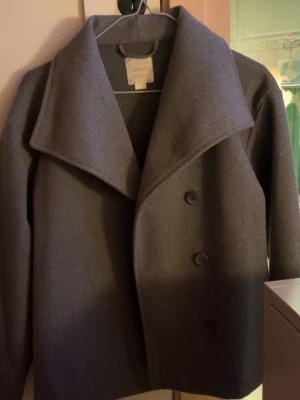 Mörkgrå ullkappa med stora slag - Stilren mörkgrå kappa med stora slag och tre knappar framtill. Klassisk och enkel design i ullmaterial, perfekt för kyligare dagar. Långärmad och med rak passform som ger en snygg siluett.