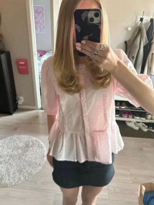 Ljusrosa volangblus med knappar - Supersöt ljusrosa blus med volang nertill och knappar framtill. Blusen har korta ärmar och ett diskret prickigt mönster i tyget. Perfekt för dig som gillar en romantisk och feminin stil.