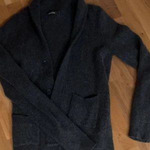 Mörkgrå kofta från Dressmann - Mysig mörkgrå kofta från Dressmann med sjalkrage och knäppning framtill. Två stora fickor framtill och ribbade muddar. Perfekt för lager på lager och enkel att matcha med olika outfits.