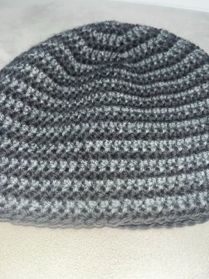 Gråmelerad virkad mössa - Säljer en gråmelerad virkad mössa med rundad form. Mössan har ett randigt mönster i olika nyanser av grått och är tillverkad med akryl garn. Perfekt för dig som vill ha en enkel men snygg Y2K accessoar till höst och vinter.