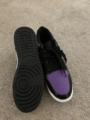 Nike Air Jordan 1 Low svart/lila/vit - Säljer ett par Nike Air Jordan 1 Low med svart, lila och vit färgkombination. Skorna har klassisk snörning, svart swoosh och vit sula. Ovandelen är i skinn med glansiga svarta detaljer och perforerad tåbox för extra stilpoäng.