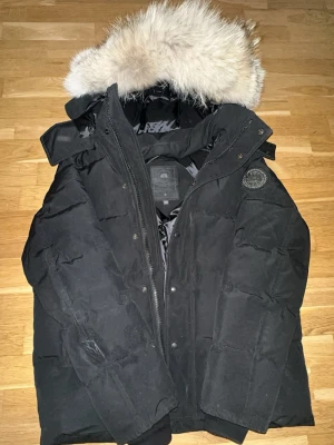  Canada Goose wyndham - Säljer en svart dunjacka från Canada Goose med stor fluffig pälskant på huvan och quiltad design. Jackan har lång ärm, dragkedja och tryckknappar framtill samt en patch på ärmen. Perfekt för kalla vinterdagar.