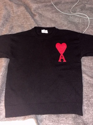Svart stickad tröja från Ami Paris - Svart stickad tröja från Ami Paris med röd hjärtlogga och bokstaven A på bröstet. Tröjan har rund halsringning och ribbade muddar vid ärmslut och nederkant. Perfekt för dig som gillar stilrena plagg med en twist.