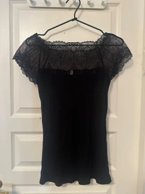 Svart topp med spetsdetaljer - Supersnygg svart topp med spetsärmar. Toppen har en figurnära passform och är gjord i ett mjukt, stretchigt material. Spetsen upptill ger en elegant och trendig look. Perfekt att styla med jeans eller kjol.