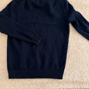 Mörkblå stickad tröja Polo Ralph Lauren - Klassisk mörkblå stickad tröja från Polo Ralph Lauren. Tröjan har rund halsringning, ribbstickade muddar vid ärmslut och nederkant samt en enkel, stilren design. Perfekt för lager på lager och passar till det mesta.