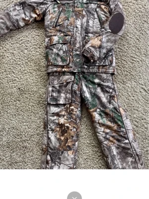 Kamo set från Swedteam med huva - Säljer ett tvådelat kamouflage set från Swedteam, perfekt för jakt, skidor eller outdoor. Setet består av jacka med huva och flera fickor samt matchande byxor. Färgen är grönbrun med löv- och grenmönster, materialet är slitstarkt och vindtätt syntet. Extra förstärkningar på armbågar och axlar.