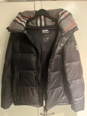 Svart Burberry dunjacka - Snygg svart dunjacka från Burberry med quiltad design och avtagbar huva. Jackan har dragkedja framtill, två sidofickor med dragkedja och en grå, rutig insida på kragen. Perfekt för kalla dagar och riktigt skön att bära.