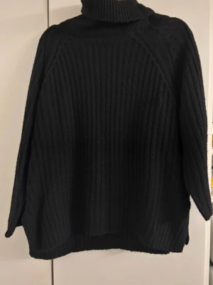 Svart ribbstickad polotröja - Mysig svart stickad polotröja med bred ribbstickning och långa ärmar. Tröjan har en hög polokrage och avslappnad passform, perfekt för kalla dagar. Materialet ser ut att vara mjukt och värmande, troligen ull- eller akrylblandning.