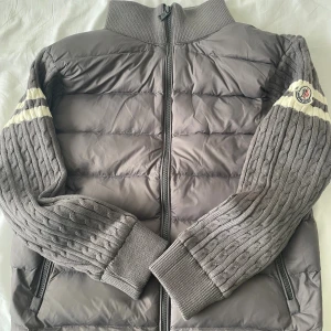 Moncler cardigan  - Snygg grå pufferjacka från Moncler med stickade kabelmönstrade ärmar och vita ränder. Jackan har hög krage, dragkedja framtill och två fickor med dragkedja. Moncler-logga på ärmen ger en exklusiv vibe. Perfekt för kyliga dagar.