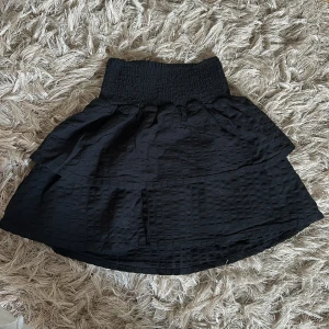 Svart volangkjol från Cope - Supersöt svart kortkjol från Cope med bred smock i midjan och volangig design. Kjolen har en diskret struktur i tyget som ger extra känsla och är perfekt för dig som gillar en enkel men ändå cool look.