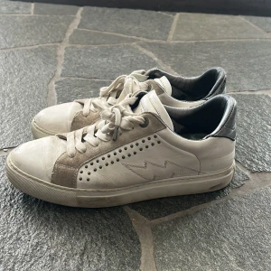 Zadig & Voltaire vita sneakers i skinn - Snygga vita sneakers från Zadig & Voltaire i äkta skinn med silvriga detaljer på hälen och insidan. Skorna har snörning, dekorativa nitar på sidorna och en blixtdetalj i sömmen. Beige mockadetaljer längs snörningen ger extra stilpoäng.