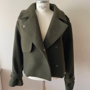 Olivgrön höstjacka från hm - Stilren olivgrön höstkappa från H&M med bred krage, dubbelknäppning och snygga spänndetaljer vid ärmsluten. Jackan är i ullblandning, passform som ger en cool vibe. Perfekt för dig som vill ha en klassisk men trendig look. Aldrig använd!