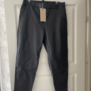 Svarta kostymbyxor i syntet - Stilrena svarta kostymbyxor från Freequent med elastisk midja och hällor för bälte. Byxorna har raka ben, snedställda fickor fram och passpoalfickor bak. Materialet har en lätt glansig finish som ger en modern look.