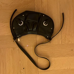 Svart axelväska i skinn med detaljer - Cool svart axelväska i skinn med unik halvmåneform, silverfärgade metalldetaljer och justerbar rem. Väskan har dragkedja och snygga spännen framtill som ger en edgy vibe. Perfekt för dig som vill sticka ut med en stilren men ändå annorlunda accessoar.