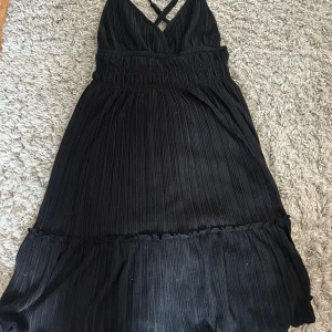 Svart omlottklänning med smala axelband - Supersnygg svart klänning med omlottliv och smala korsade axelband i ryggen. Klänningen har plisserat tyg, markerad midja och volangkant nertill. 