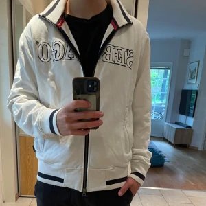 Vit Sebago fullzip kofta med brodyr - Säljer en vit ziptröja från Sebago med broderad logga över bröstet. Tröjan har svarta detaljer vid krage, ärmslut och midja samt dragkedja framtill. Skön passform och perfekt för en clean och sportig look då det är ett seglar märke. Tröja passade för höst och vinter