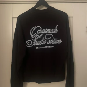 Svart original från Jack & Jones - Svart original-tröja från Jack & Jones med ljusblå text 'Originals Studio Edition' på ryggen och mindre tryck på bröstet. Klassisk passform med rund hals och långa ärmar. Perfekt för dig som vill ha en enkel och skön overshirt| Skriv vid minsta fråga elle fundering| pris ej hugget i sten 