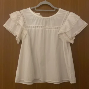 Vit blus med volang från Vero Moda - Supersöt vit blus från Vero Moda i bomull med volangärmar och spetsdetaljer längs axlar och ärmslut. Blusen har rund halsringning och knapp i nacken. Perfekt för en fräsch och luftig look under varmare dagar.