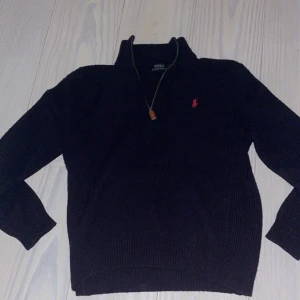 Svart stickad tröja Polo Ralph Lauren Storlek 167-173 - Svart stickad tröja från Polo Ralph Lauren med halv dragkedja och klassisk röd logga broderad på bröstet. Tröjan har hög krage och långa ärmar, perfekt för kyligare dagar. Snygg och tidlös modell som passar till det mesta.