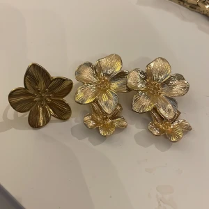 Guldiga blommiga örhängen och ring - Säljer ett set med stora, blommiga smycken i guldmetall – ett par örhängen med tre blommor i olika storlekar och en matchande ring med en blomma. Perfekt för dig som vill sticka ut med ett unikt och trendigt statement.