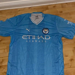 Manchester City Haaland matchtröja Puma - Säljer en officiell Manchester City fotbollströja från Puma med Haaland och nummer 9 på ryggen. Tröjan är ljusblå med tunna ränder, klubbmärke på bröstet och Etihad Airways som sponsor. Korta ärmar och tillverkad i lätt, ventilerande material.