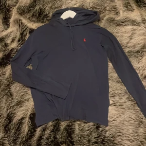 Mörkblå hoodie från Polo Ralph Lauren - Mörkblå hoodie från Polo Ralph Lauren med röd broderad logga på bröstet. Tröjan har luva med snörning och är tillverkad i bomull. Passar till allt och är även skön att ha på sig när man bara ska chilla. Det är köpt helt ny på nätet och gör för cirka 1200kr. Jag säljer tyvärr nu för att den är påväg att bli för liten. Finns inga defekter på den. Skriv vid fundering. 