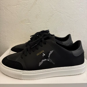 Svarta sneakers från Axel Arigato - Snygga svarta sneakers från Axel Arigato med vit sula och svart snörning. Skorna har en broderad fågel på sidan och guldfärgad Arigato-text. Tillverkade i skinn med detaljer i mocka och grå hälkappa. Perfekta för en clean och stilren look. Nypris 2750 kr. Enbart använda en gång!
