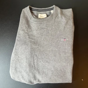 Grå stickad tröja från GANT - Säljer en stilren grå stickad tröja från GANT med broderad logga på bröstet. Tröjan har rund halsringning med ribbade detaljer och passar perfekt till lager-på-lager-stil. Enkel att matcha med jeans eller chinos.