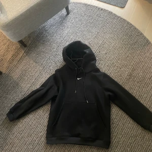 Svart Nike hoodie med huva och ficka - Svart hoodie från Nike med klassisk vit swoosh-logga på bröstet. Tröjan har huva med snörning och en stor magficka. Tillverkad i mjuk bomullsblandning och har långa ärmar. Perfekt för en chill och sportig stil. Helt oanvänd 