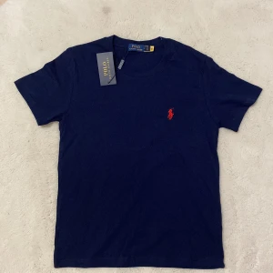 Marinblå t-shirt från Polo Ralph Lauren - Snygg marinblå t-shirt från Polo Ralph Lauren i mjuk bomull. Klassisk passform med korta ärmar och rund hals. Ikonisk röd broderad logga på bröstet. Perfekt basic med stilren vibe.