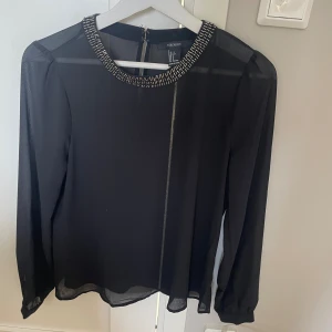 Svart blus med pärldetaljer Forever 21 - Snygg svart blus från Forever 21 med transparenta långärmar och rund halsringning dekorerad med silvriga pärlor. Blusen är lätt och luftig, perfekt för att lyfta din outfit med lite extra bling. Stängs med dragkedja bak.