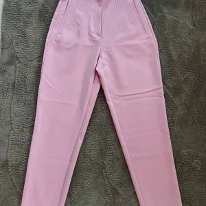 Rosa högmidjade byxor från Zara XS - Snygga ljusrosa byxor från Zara i storlek XS. Byxorna är högmidjade med raka pressveck framtill, har två fickor och är tillverkade i ett slätt, vävt material. Perfekta för en trendig och stilren look.