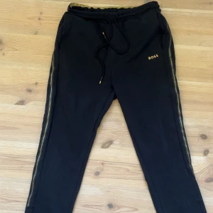 Svarta mjukisbyxor från BOSS med guld - Svarta mjukisbyxor från BOSS med guldfärgade detaljer och logga på vänster ben. Byxorna har resår och snörning i midjan samt guldfärgade ränder längs sidorna. Perfekta för en chill och snygg look.