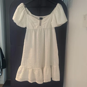 Vit kortklänning från Gina Tricot - Supersöt vit kortklänning från Gina Tricot med offshoulder-ärmar och volang nertill. Klänningen har en strukturerad, lite krinklad yta och en snygg v-ringning framtill. Perfekt för varma dagar och somriga vibes.