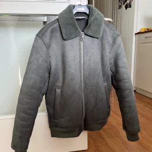 Grå teddyjacka med dragkedja - Snygg grå teddyjacka med mjuk, fluffig krage och hel dragkedja framtill. Jackan har ribbade muddar vid ärmslut och midja samt två diskreta fickor på sidorna. Perfekt för dig som gillar stilren och enkel design med en cozy touch.