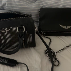 Två svarta Zadig & Voltaire väskor - Säljer två svarta väskor från Zadig & Voltaire i skinn. Ena är en handväska med silvernitar, dubbla handtag och dragkedjor. Den andra är en axelväska med kedjerem och silvervinge framtill. Båda har coola detaljer i silver och passar perfekt till en edgy look. Den ena e en limetedichen och den har används fler tal och den andra e helt oanvänd. Köpta i Schweiz i Zürich❤️