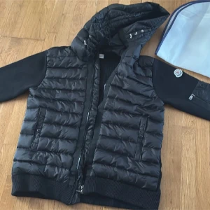 Moncler cardigan  - Svart pufferjacka från Moncler med stickade ärmar och quiltad kropp. Jackan har huva med snörning, dragkedja framtill och fickor med dragkedja. På ena ärmen finns en ficka med dragkedja och Moncler-logga. Materialmix av polyester och stickat ger en modern look.