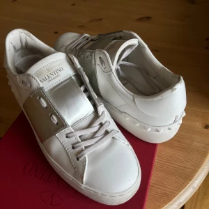 Valentino Garavani Open sneakers vit/silver - Snygga Valentino Garavani Open sneakers i vitt skinn med en bred metallic-silver detalj över vristen. Klassisk rund tå, platt sula och coola nitar baktill. Skorna har snörning och kommer med en originalkartong och dustbag! Skorna har genomgått en äkthetskontroll genom Vestiare, skorna har även koden kvar på tungans undersida. Priset kan diskuteras, Hör av dig vid frågor!!💗
