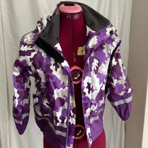 Lila galonjacka från PAX, strl 130 - Galonjacka från PAX i lila, vit och beige camouflagemönster. Jackan har löstagbar huva, reflexdetaljer och dragkedja framtill. Insidan är fodrad med mjuk fleece vid kragen för extra komfort. Kan inte komma ihåg att denna jacka någonsin använts så den är i bra skick förutom ett litet  hål (se bild 4&5) som går att tejpa.