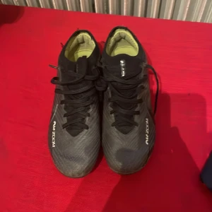 Nike Air Zoom svarta fotbollsskor - Säljer ett par svarta Nike Air Zoom fotbollsskor med snörning och låg profil. Skorna har en texturerad ovandel i syntetmaterial och gula detaljer på insidan och sulan. Perfekta för dig som vill ha grymt grepp och stil på planen.