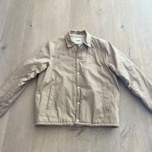 Beige teddyjacka från Dobber - Snygg beige teddyjacka från Dobber med mjukt, vitt teddyfoder på insidan. Jackan har klassisk krage, raka fickor, knappar framtill och dragsko i nederkant. Perfekt för kyliga dagar och enkel att matcha med det mesta.