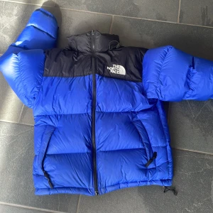 Blå och svart pufferjacka The North Face - Säljer en blå och svart pufferjacka från The North Face. Jackan har hög krage, dragkedja framtill och två fickor med dragkedja. Klassisk logga på bröstet och ryggen. Perfekt för kalla dagar när du vill hålla värmen och ändå se stilren ut.Har litte fläckar men inget som man tänker på har hafft den i en säsong men börjar bli för litte nu priset är inte ristat i sten 