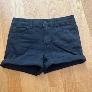 Svarta Cubus shorts med upprullad kant - Svarta shorts från Cubus i klassisk femficksmodell med upprullad kant nertill. De har bälteshällor, knapp och dragkedja framtill. Perfekta för sommaren och enkla att matcha med allt. Materialet känns mjukt och stretchigt.