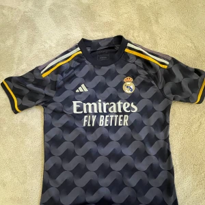 Real Madrid bortatröja Vini Jr. 7 Adidas - Säljer en mörkblå Real Madrid bortatröja från Adidas med Vini Jr. och nummer 7 på ryggen. Tröjan har korta ärmar, gula detaljer och ett vågigt mönster. Broderat klubbmärke på bröstet och tryckt sponsorlogga framtill. Tillverkad i lätt och ventilerande material. Köpt från unisport. Lite av trycket på S:et har åkt av på emirates märket.