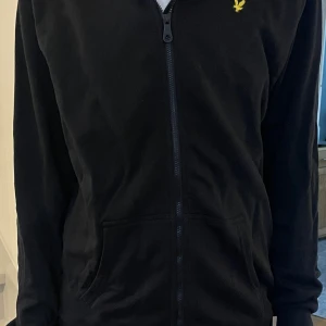 Svart hoodie från Lyle & Scott - Svart hoodie från Lyle & Scott med dragkedja framtill och klassisk huva. Tröjan har en gul broderad logga på bröstet och praktiska fickor framtill. Perfekt för chill dagar.