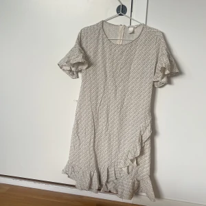 Beige mönstrad volangklänning - Supersöt beige klänning med diskret mönster, volangdetaljer nertill och korta ärmar. Klänningen har rund halsringning och lätt struktur i tyget, perfekt för en avslappnad men ändå stilsäker look.