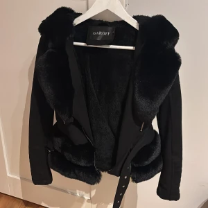 Garoff pälsjacka med luva - Svart pälsjacka från Garoff med stor fluffig huva. Jackan har bälte i midjan. Storlek S men passar mig som har XS annars. Finns i Stockholm.